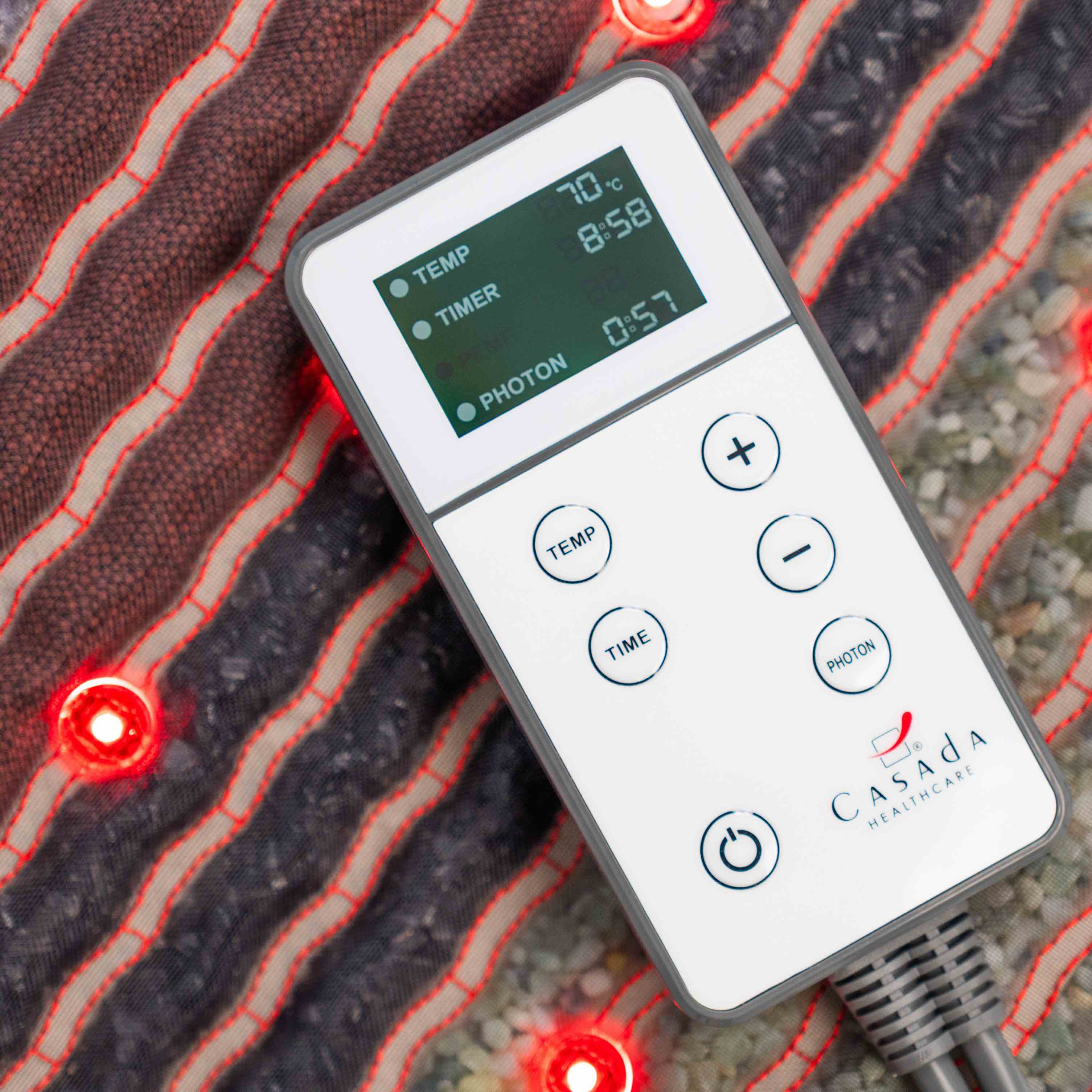 Pilot sterujący Casada z czytelnym wyświetlaczem LCD do regulacji temperatury, czasu i trybu fotonowego, położony na podgrzewanej macie masującej z czerwonymi diodami LED.