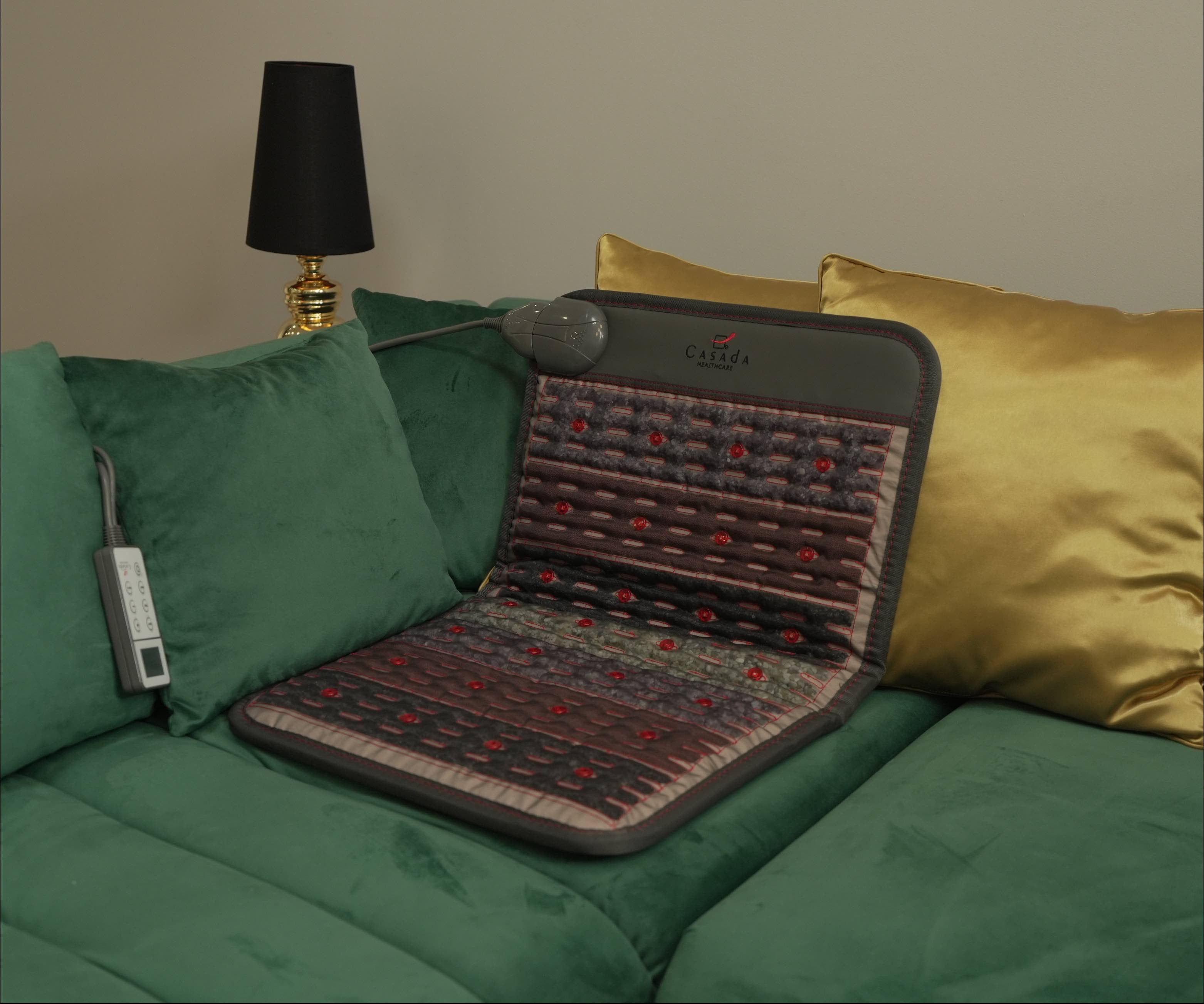 Podgrzewana mata masująca Casada ułożona na zielonej sofie, z widocznymi czerwonymi diodami LED i pilotem sterującym, prezentująca zastosowanie urządzenia w domowym wnętrzu.