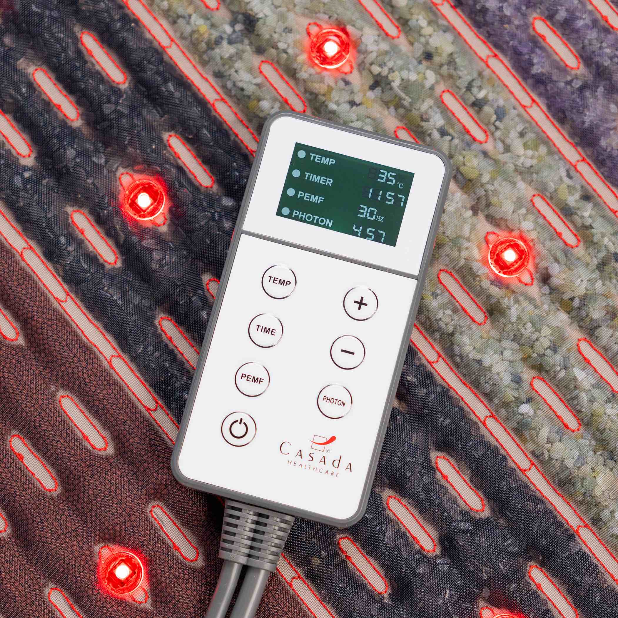 Pilot sterujący Casada z czytelnym wyświetlaczem LCD do regulacji temperatury, czasu i trybu fotonowego, położony na podgrzewanej macie masującej z czerwonymi diodami LED.