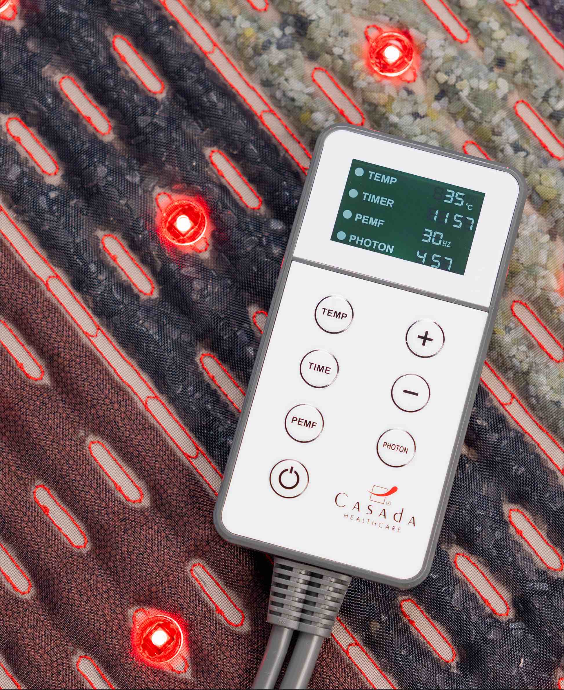Pilot sterujący Casada z czytelnym wyświetlaczem LCD do regulacji temperatury, czasu i trybu fotonowego, położony na podgrzewanej macie masującej z czerwonymi diodami LED.