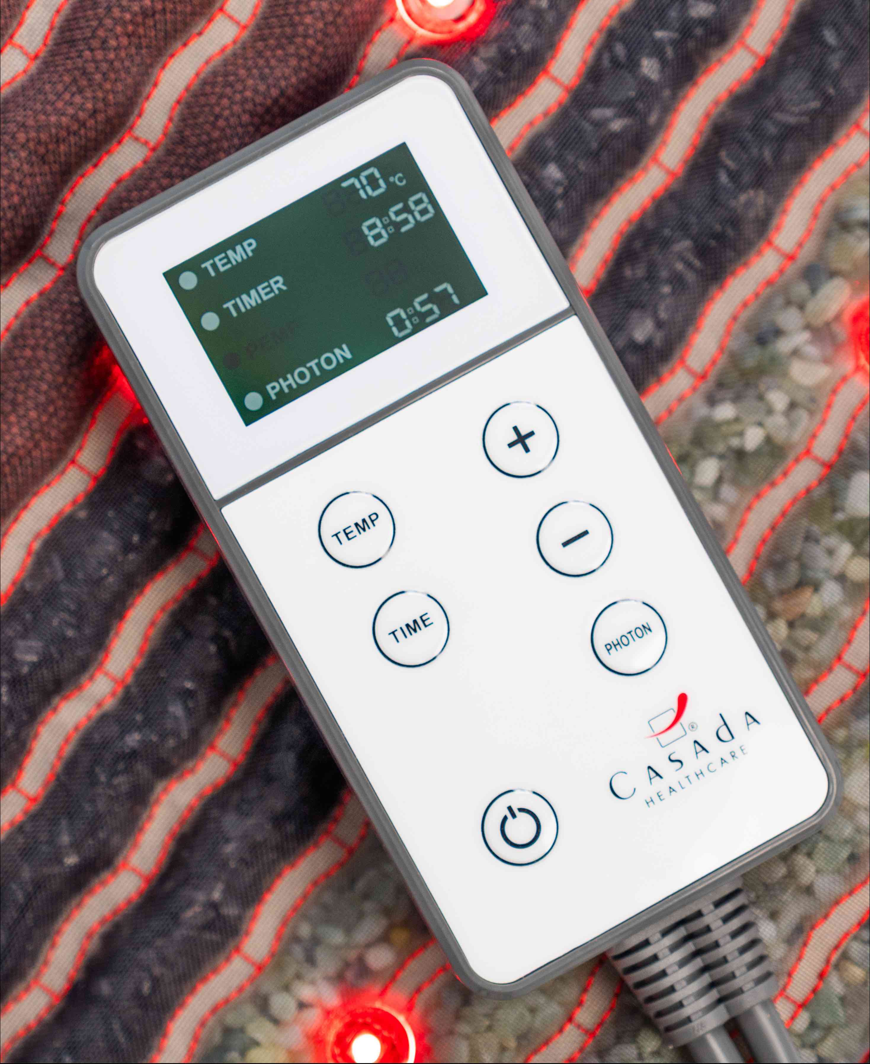 Pilot sterujący Casada z wyświetlaczem LCD pokazującym temperaturę, czas i tryb fotonowy, umieszczony na podgrzewanej macie masującej z czerwonymi diodami LED.