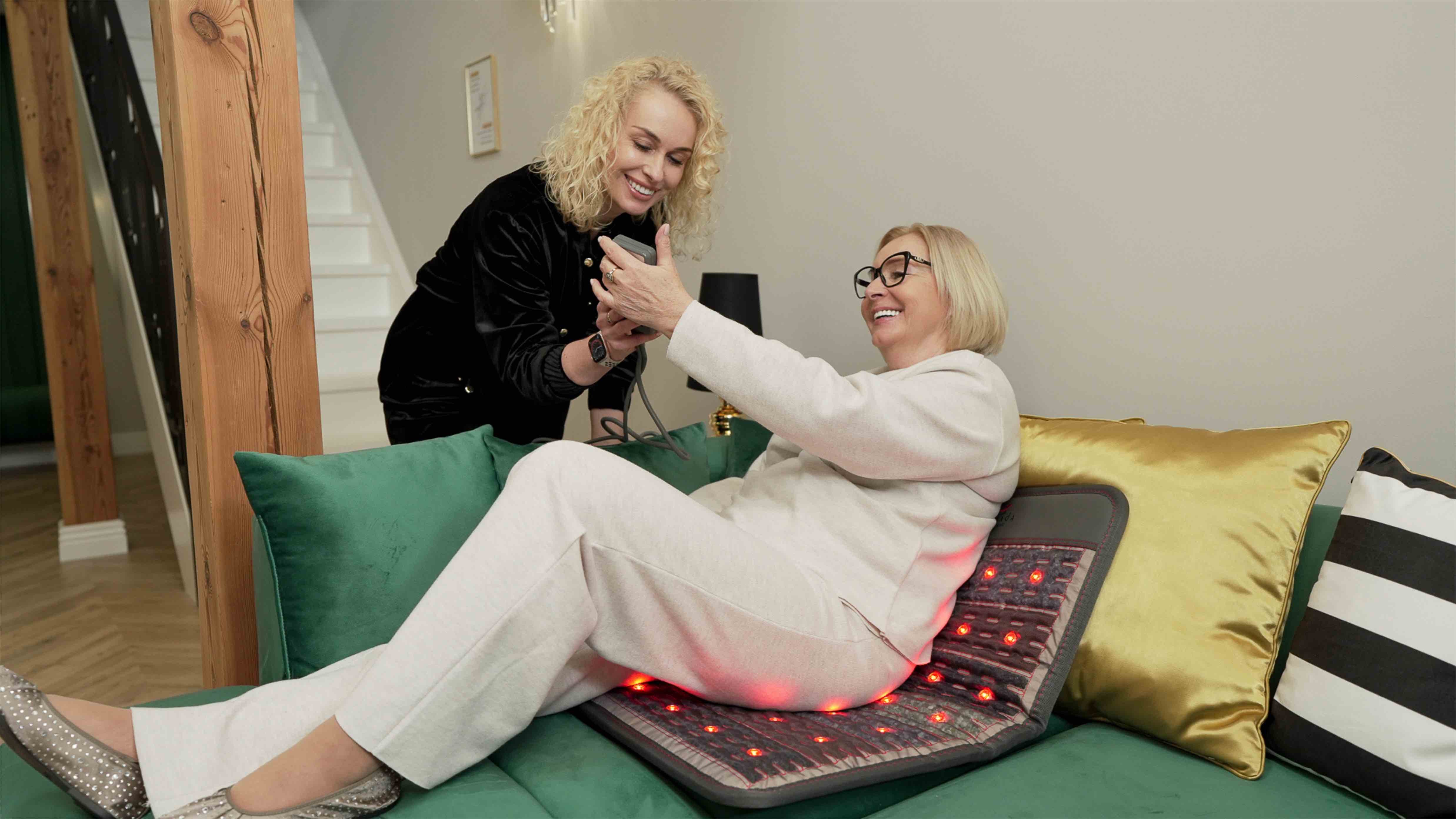 Starsza kobieta siedzi na włączonej macie grzejącej Dr. Stone PRO, a młodsza prezentuje jej działanie pilota.