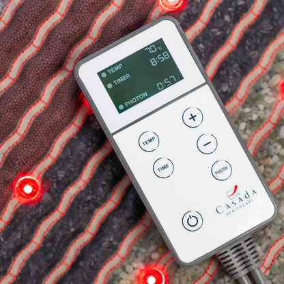 Pilot sterujący Casada z czytelnym wyświetlaczem LCD do regulacji temperatury, czasu i trybu fotonowego, położony na podgrzewanej macie masującej z czerwonymi diodami LED.