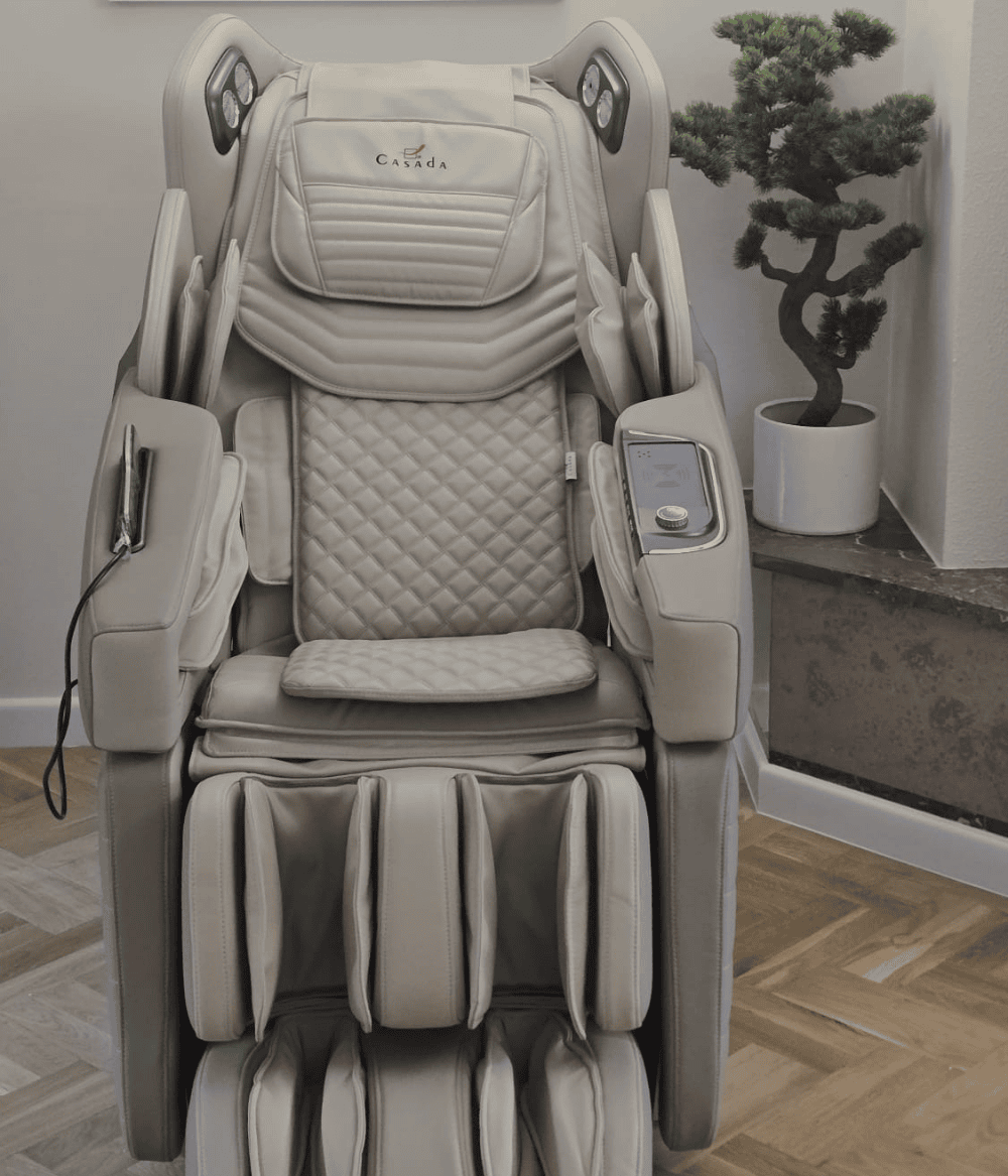 Jasny fotel masujący z pikowanym siedziskiem i panelem sterowania na podłokietniku, ustawiony we wnętrzu z drewnianą podłogą i rośliną w donicy